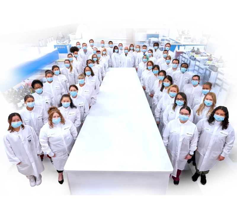 Laboratory employees in white coats standing around a long white table. ACS Laboratory.